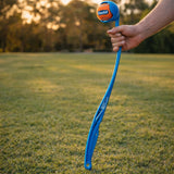 Hand holding a long blue Chuckit ball launcher with orange ball in a park
