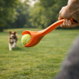 Person holding an orange ball launcher with a tennis ball, dog running in the background